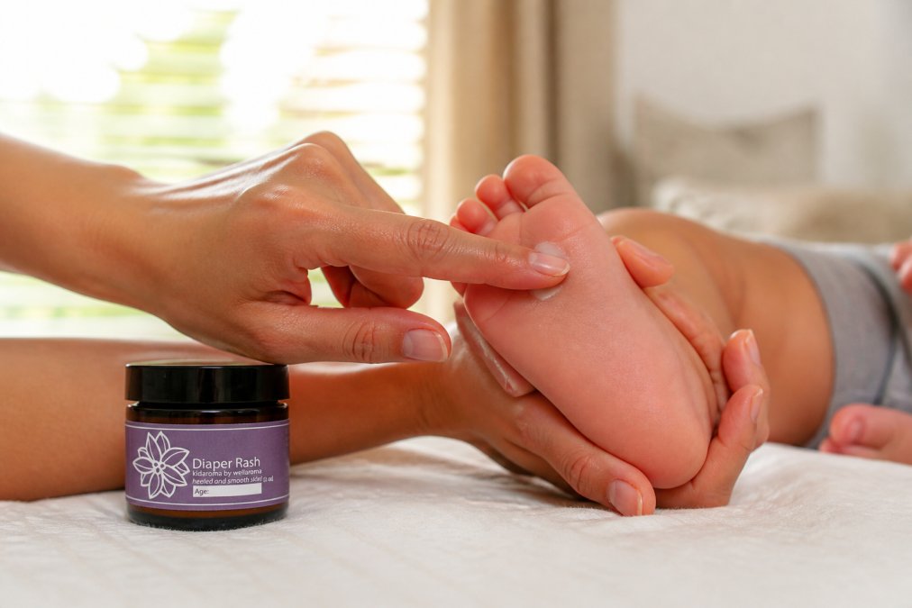 Parent's hand applying a small pinch of diluted essential oil cream to a child's foot using fingertip, showing gentle topical application with natural sunlight in background