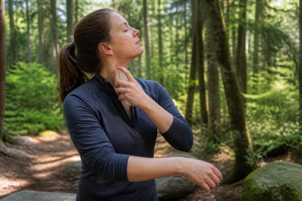 Person in a wooded outdoor setting applying insect repellent to their neck and exposed arms, wearing long sleeves pushed up, with dappled sunlight filtering through trees