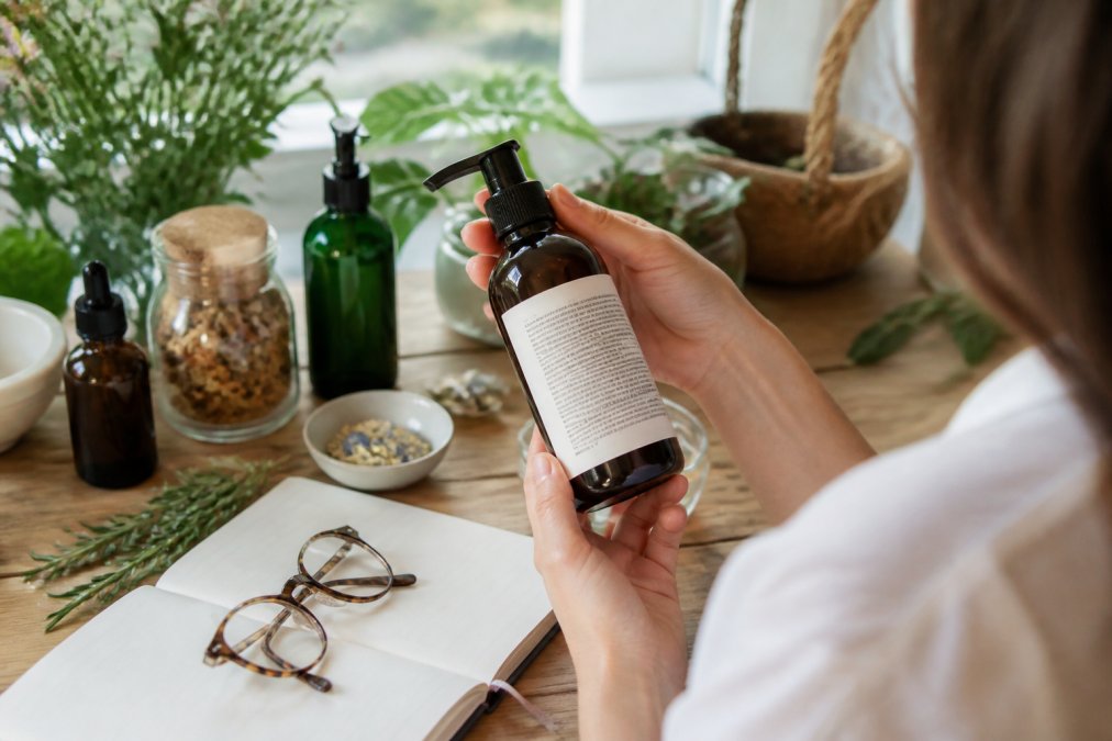 Person's hand holding a premium hair care product bottle, carefully examining the detailed ingredient label in natural window lighting, with reading glasses nearby