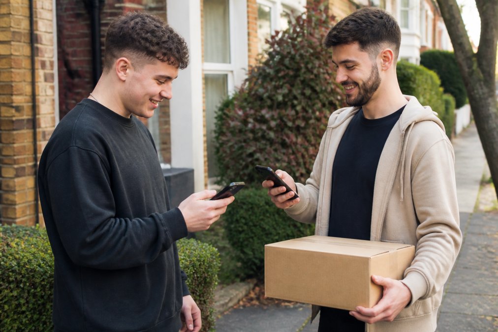 Two people using mobile phones to verify safe payment methods while meeting outdoors to exchange a boxed item
