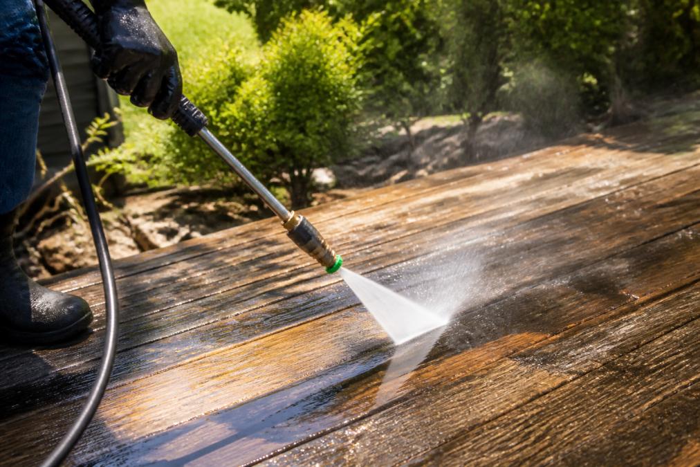 A gloved hand holding a pressure washer wand at a low angle, spraying a wide fan of water across weathered timber deck boards in bright afternoon sunlight
