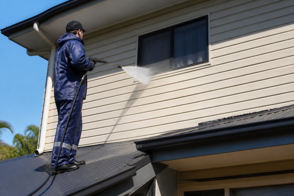 Professional cleaner in protective gear using a pressure washer wand with a wide fan spray to rinse vinyl siding on a two-storey Australian home, working from the top down in bright daylight