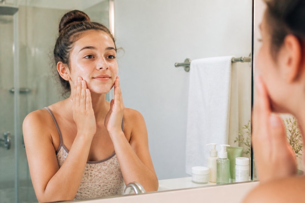 A teen girl with a calm, clear expression applying a gentle cream cleanser to her face in bright natural bathroom lighting, with skincare products neatly arranged on the counter beside her