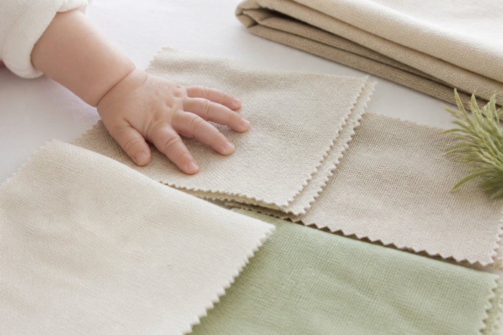 Close-up of soft, natural fabric swatches in neutral tones (organic cotton, linen, bamboo) arranged on a clean white surface with a baby's hand gently touching one swatch