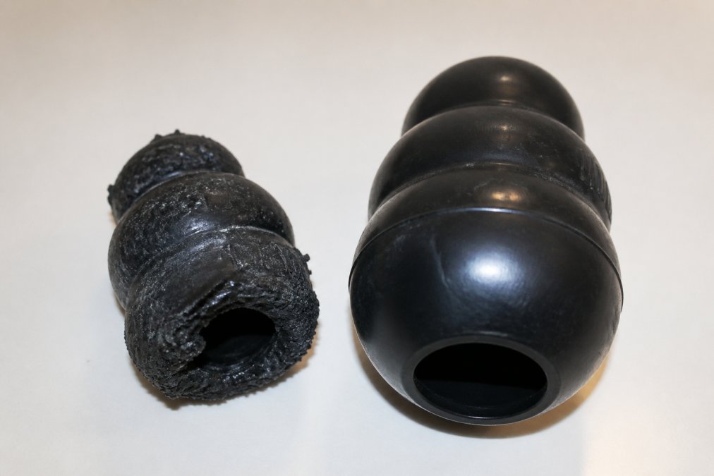 Close-up of a heavily worn, cracked black rubber dog toy placed next to an intact replacement toy on a white surface, showing visible surface crazing, torn edges, and significant size reduction on the damaged toy