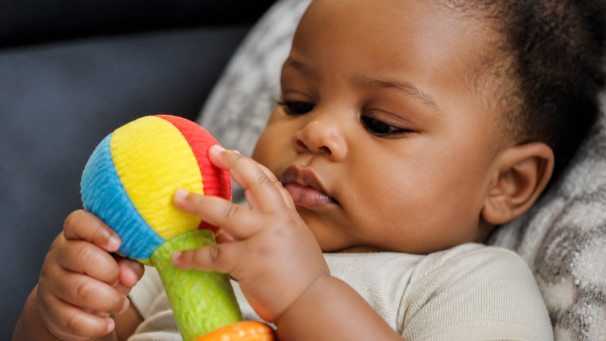 Close-up of a baby's hand grasping a soft, textured rattle with bright primary colours, showing focused concentration and finger exploration