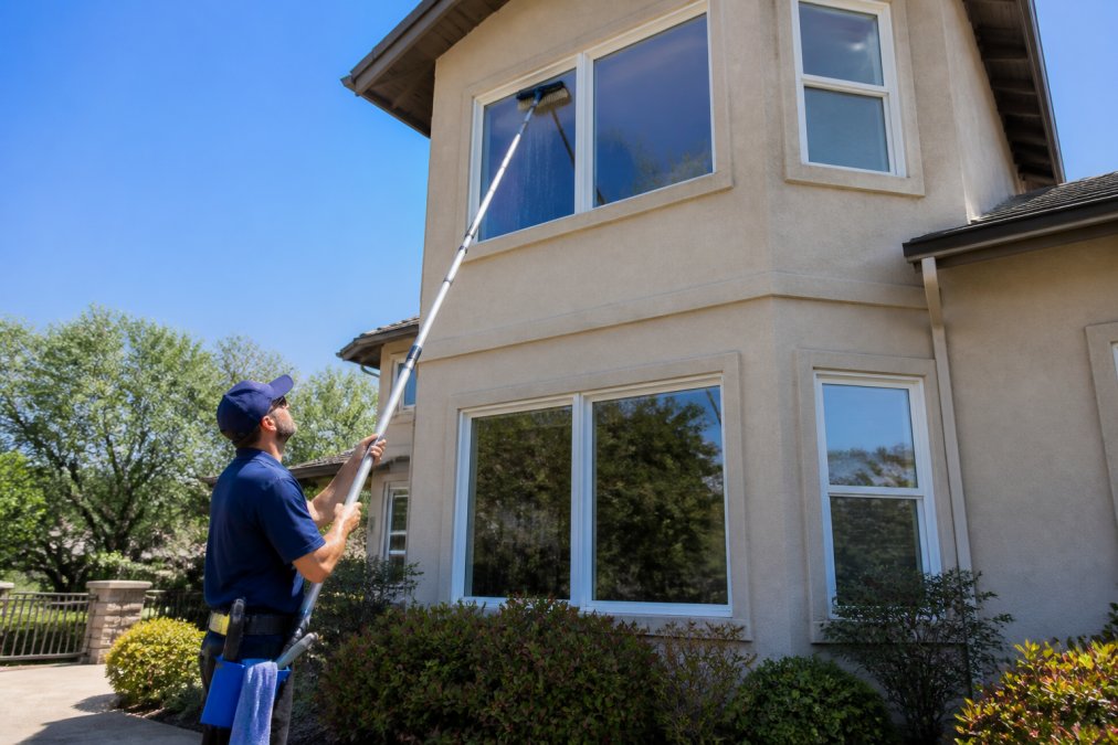 A homeowner standing on solid ground using a long extension pole with a scrubber head to clean a second-story window on a two-story house, bright daylight, no ladder visible