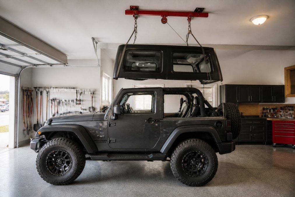 A Jeep Wrangler with its hardtop being lifted by a red hoist system in a clean, well-lit garage, hardtop suspended safely above the vehicle, clear garage floor