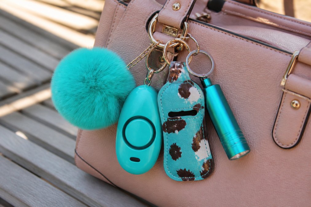 Close-up of a stylish turquoise cow print keychain set with personal alarm, mini flashlight, and pom pom accessory displayed on a designer purse with natural daylight highlighting the details