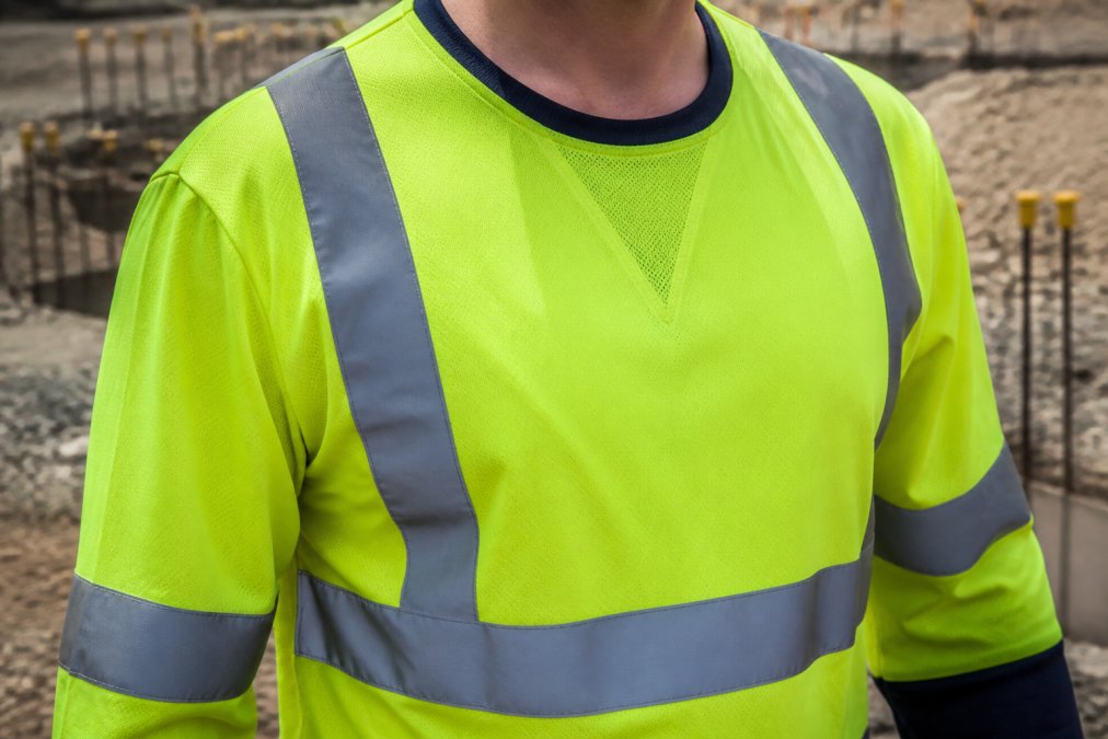 Close-up of a worker's high-visibility long-sleeve shirt showing reflective tape bands on the sleeves and body, with breathable mesh visible at the collar, worn on an outdoor construction site