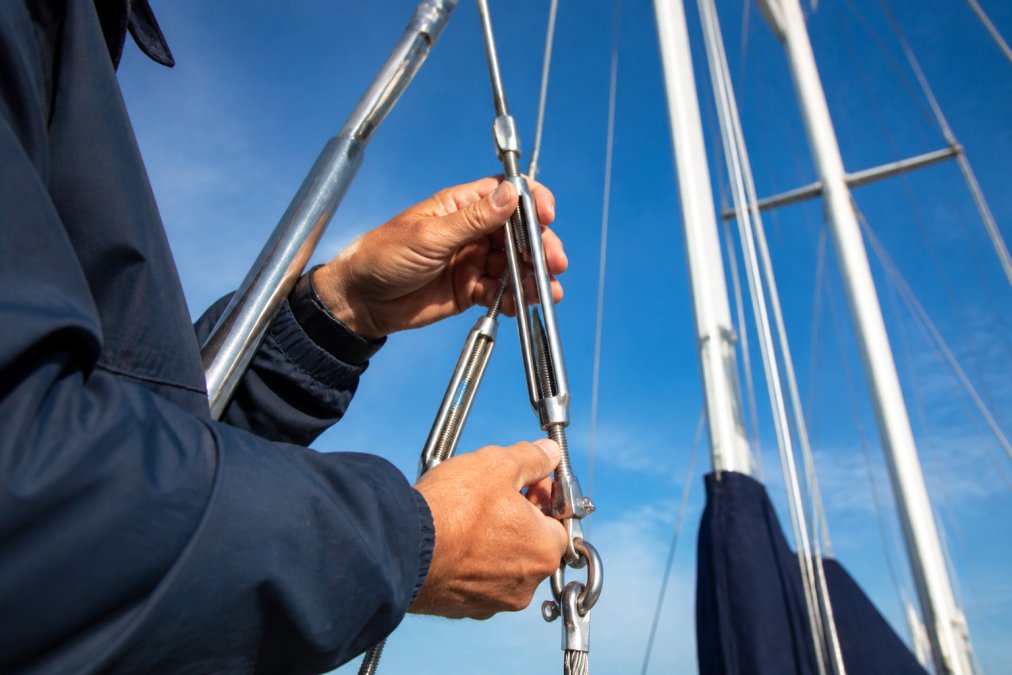 Close-up of a sailor's hands inspecting turnbuckles and swaged terminals on standing rigging, with wire rope and shackles visible against a clear sky and mast above