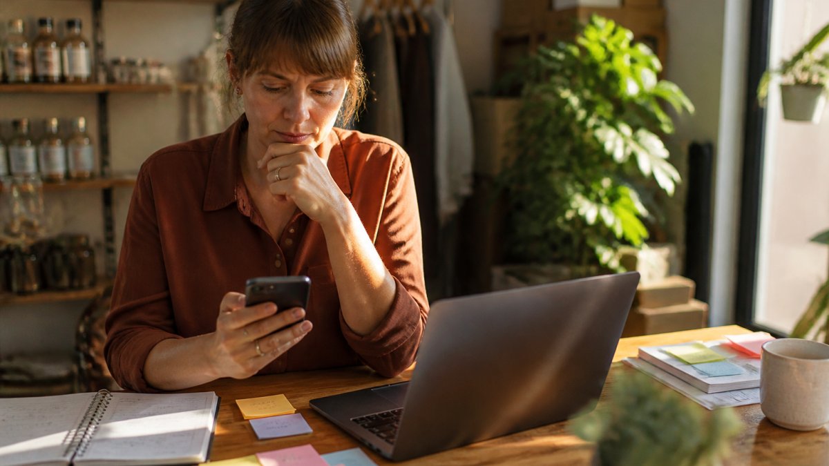Small business owner checking phone and laptop at desk surrounded by sticky notes and notebook, focused on managing multiple enquiries