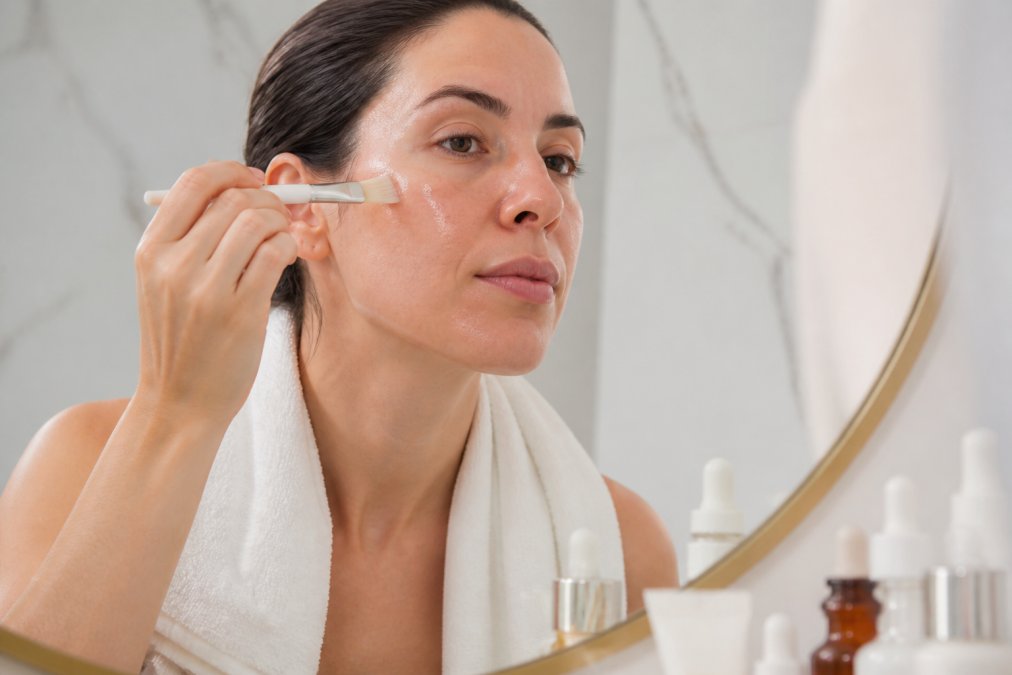 Close-up of a woman carefully applying a clear chemical peel solution to her clean face with a small brush, looking into a well-lit bathroom mirror, with a clean white towel draped over her shoulders