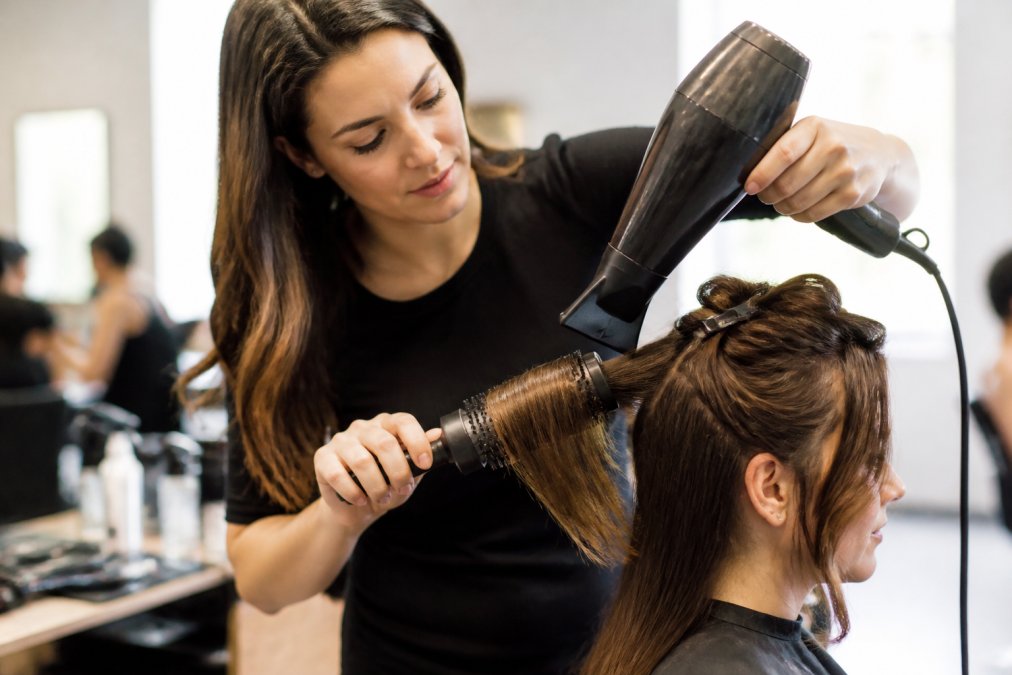 Professional stylist demonstrating round brush technique on damp hair section, using concentrator nozzle with proper airflow direction, hands positioned for tension control