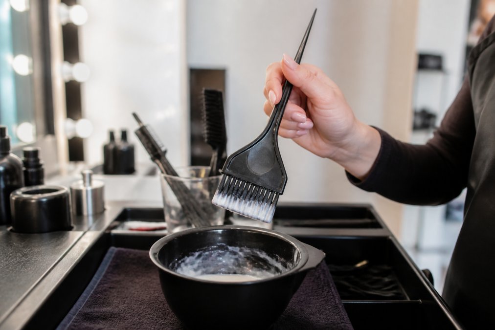 Close-up of a professional colorist's hand gripping a wide-based feathered brush over a bowl, color visible on the bristle tip, bright salon lighting with tools arranged on a clean counter