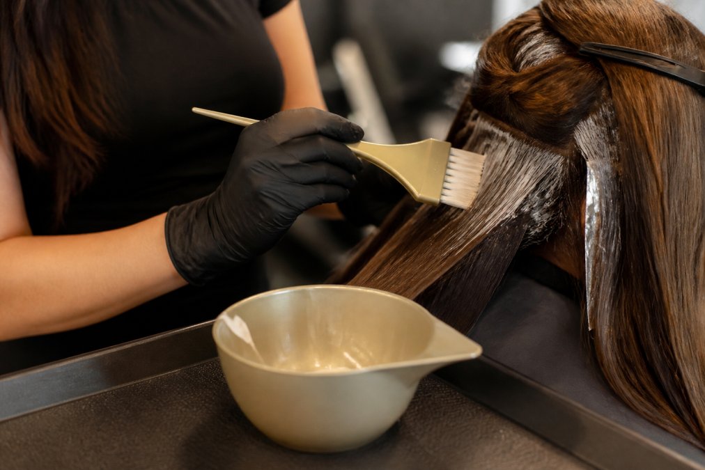 Professional hair colorist at salon station applying color with feathered brush to client's hair, showing proper brush grip and stain-resistant bowl placement on work surface with natural studio lighting