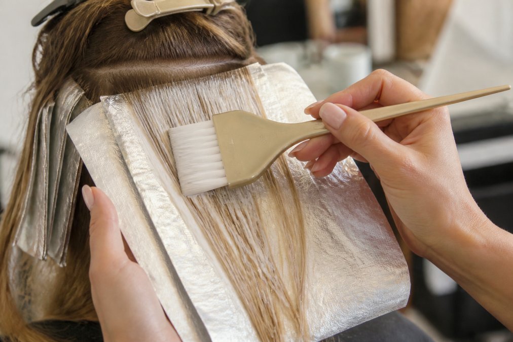 Close-up of colorist's hands using no-slip foils and precision feathered brush to apply balayage color to sectioned hair, demonstrating technique and foil stability with professional lighting