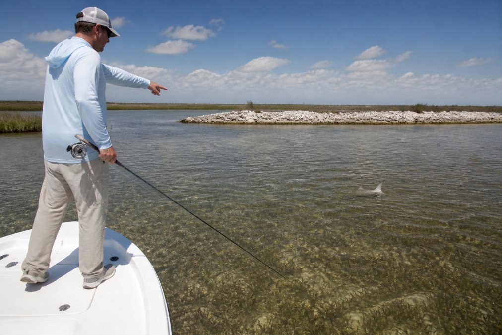 An angler standing on the bow of a shallow-water skiff, pointing toward a tailing redfish on a grass flat during a rising tide, with an oyster bar visible in the background