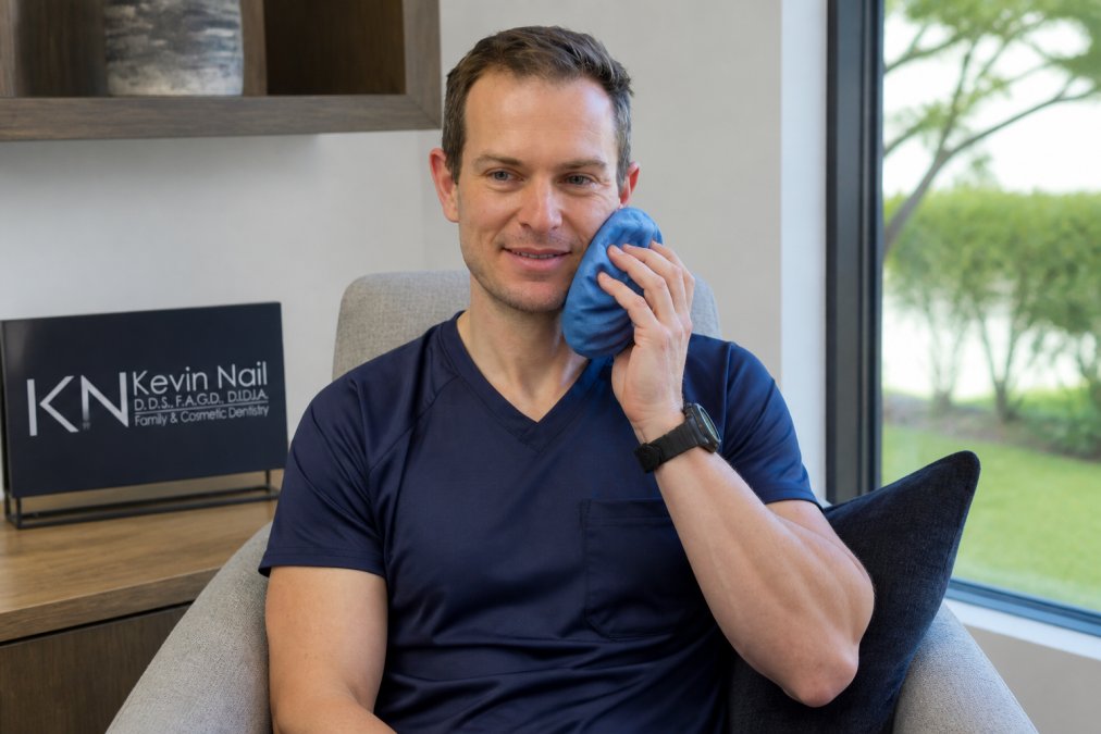 Patient holding ice pack wrapped in soft cloth to cheek while sitting in comfortable chair with natural window lighting, calm expression