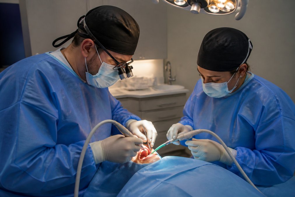 Dental professional in surgical attire performing implant placement procedure with specialized instruments and overhead surgical lighting in a modern dental surgery suite
