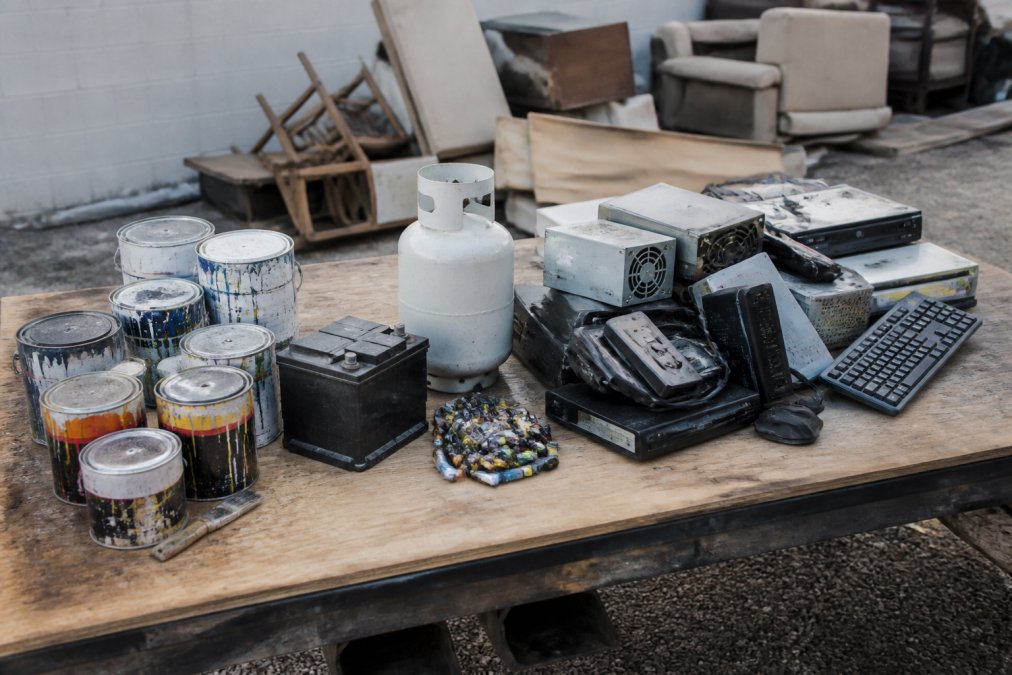 Pile of hazardous materials and restricted items including paint cans, batteries, propane tanks, and e-waste clearly separated from general household junk on a work surface