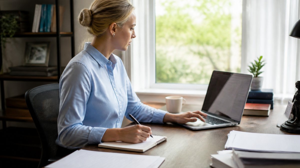Professional sitting at organized home office desk with laptop, documents, and notepad, preparing for video consultation with natural window lighting