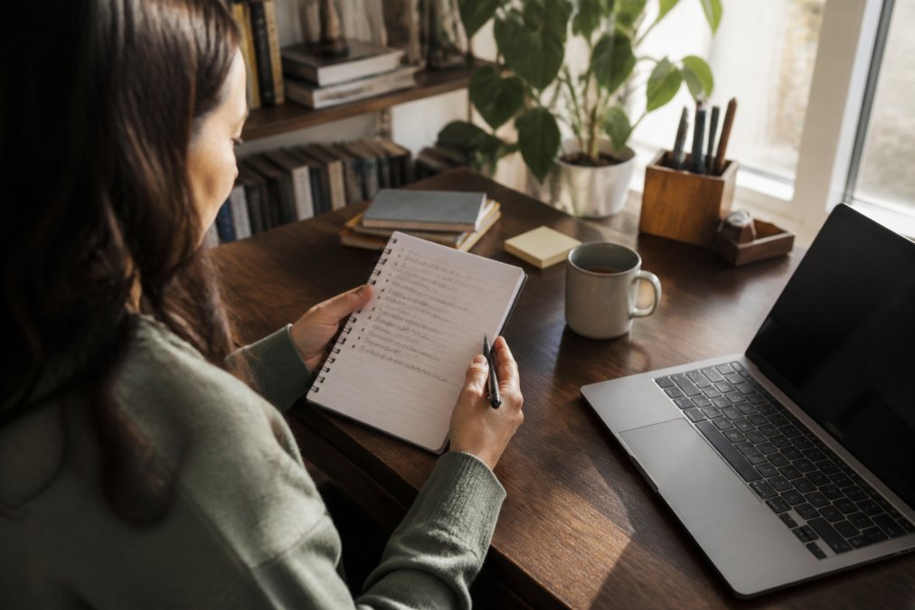 A person sitting at a desk with a notepad, pen, and laptop, reviewing a handwritten list of questions for a doctor's appointment, in a calm home office with soft afternoon light