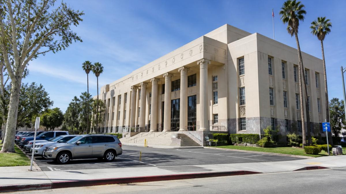 Exterior view of the San Fernando Courthouse building with parking lot visible, showing the building facade, entrance area, and surrounding street