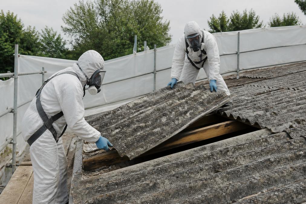 Pracovníci v ochranných bílých kombinézách s celoobličejovými respirátory provádějící demontáž azbestové střešní krytiny na stavbě s izolovanou pracovní plochou