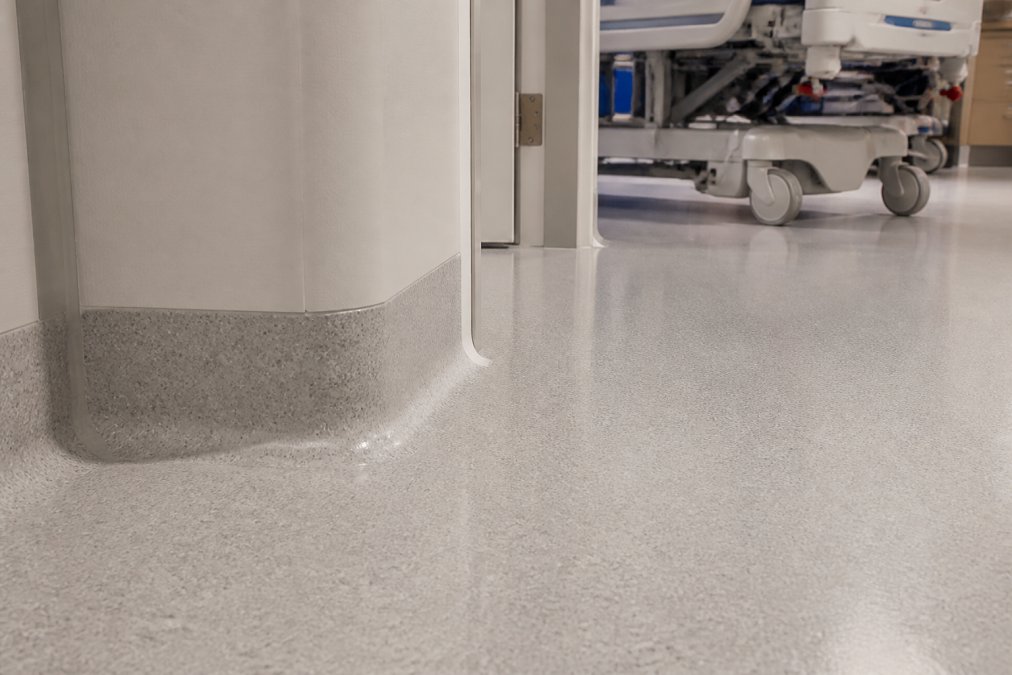 Close-up of pristine hospital flooring showing seamless welded seams meeting the wall with a coved base design, medical equipment visible in soft clinical lighting