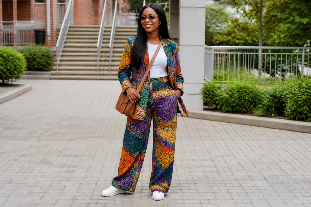 Woman wearing a vibrant multicoloured Ankara print co-ord set with matching jacket and wide-leg trousers, styled with clean white trainers and a simple crossbody bag, standing outdoors in natural daylight with confident posture
