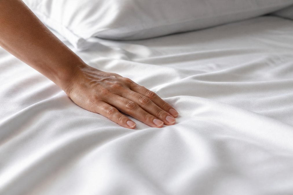 Close-up of a hand smoothing a white bamboo sheet on a bed, showing the subtle sheen and tight weave of the fabric under soft morning light