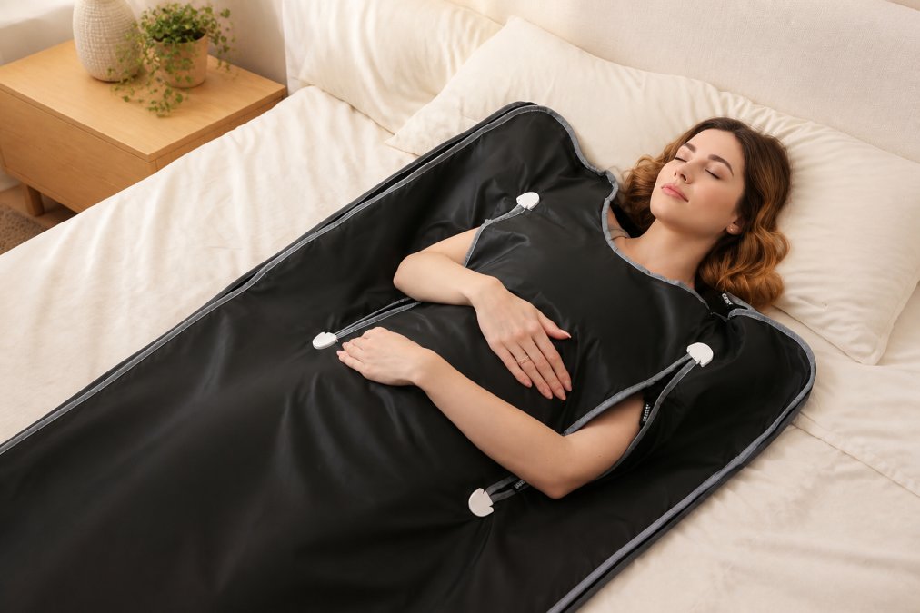 Person lying relaxed in a portable infrared sauna blanket at home with warm ambient lighting, showing the blanket wrapped around their body in a comfortable reclined position