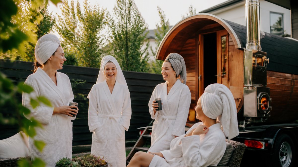 A group of women in robes and towels laughing outside a wood-fired sauna trailer in a backyard, string lights overhead, late afternoon light