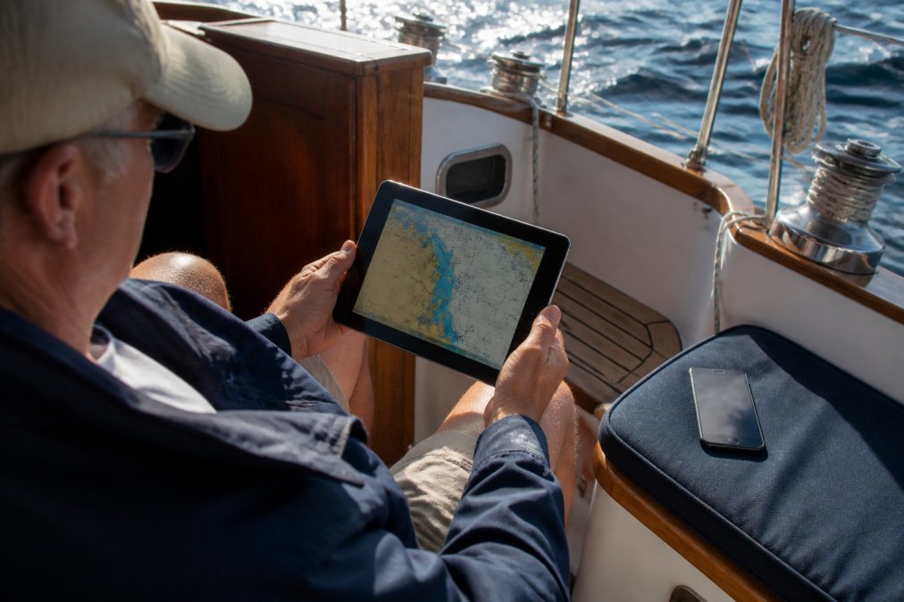 A boat owner in a sailboat cockpit checking a tablet displaying a marine chartplotter interface, with a smartphone showing a price alert notification lying on the seat beside them, bright daylight