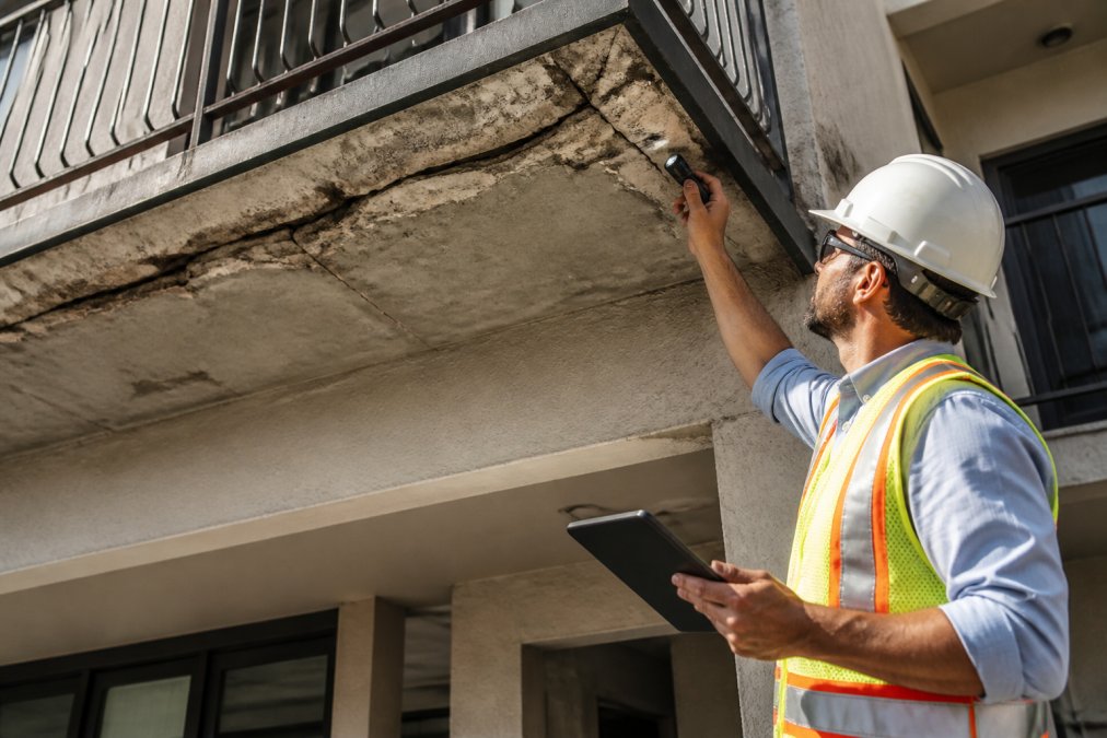 A licensed inspector in a hard hat and safety vest examining the underside of a multi-story apartment building's exterior balcony with a flashlight while documenting findings on a tablet in bright daylight