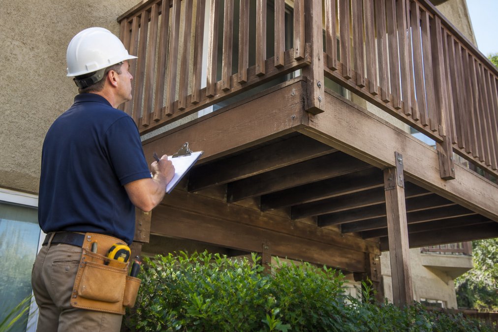 Licensed structural inspector examining a residential balcony in daylight, using a clipboard to document findings while carefully inspecting the railing connections, deck surface, and visible framing, with measurement tools visible on the work belt