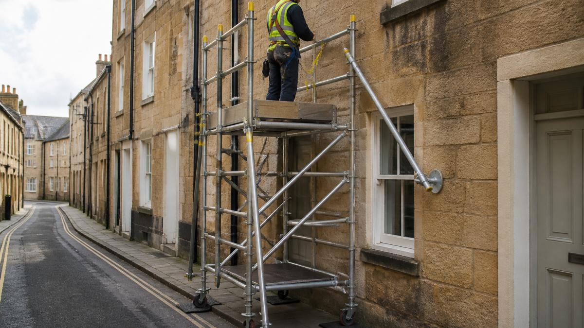Compact mobile access tower positioned against a period stone terraced building on a narrow street, showing protective rubber mats under base plates, diagonal bracing secured to stonework, and a worker on the platform with safety harness and fall protection equipment