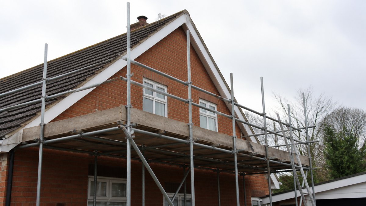 Professional scaffolding erected against residential home with pitched roof, showing horizontal working platform with safety railings providing secure access for roof repair work