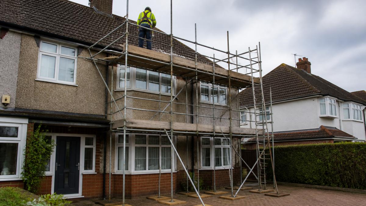 A residential semi-detached house with a two-storey scaffold tower erected along the front elevation, base plates resting on timber sole boards across a paved driveway, a roofer in a harness working on the roof edge, overcast British sky