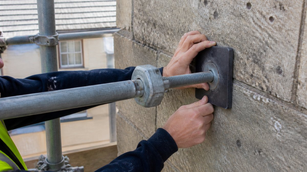 Close-up of protective rubber pads and conservation-compatible scaffold ties being carefully positioned against period Bath stone masonry, showing technician's hands adjusting placement to avoid damage