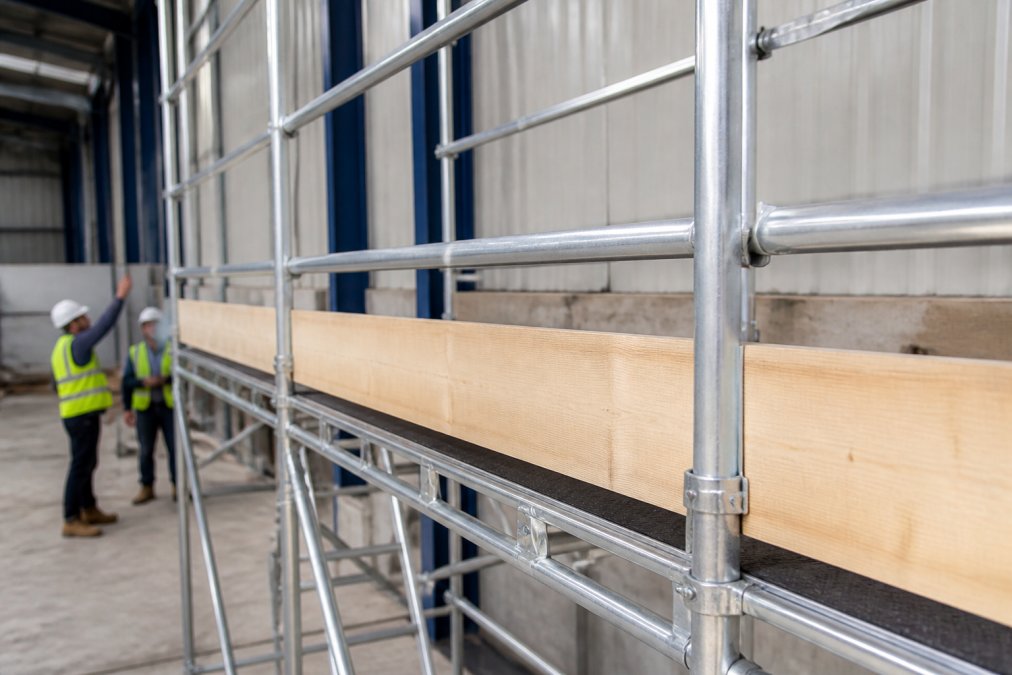 Close-up of properly installed guardrails and toe boards on an aluminium scaffold tower on a construction site, showing correct height and secure fastening with workers visible in background