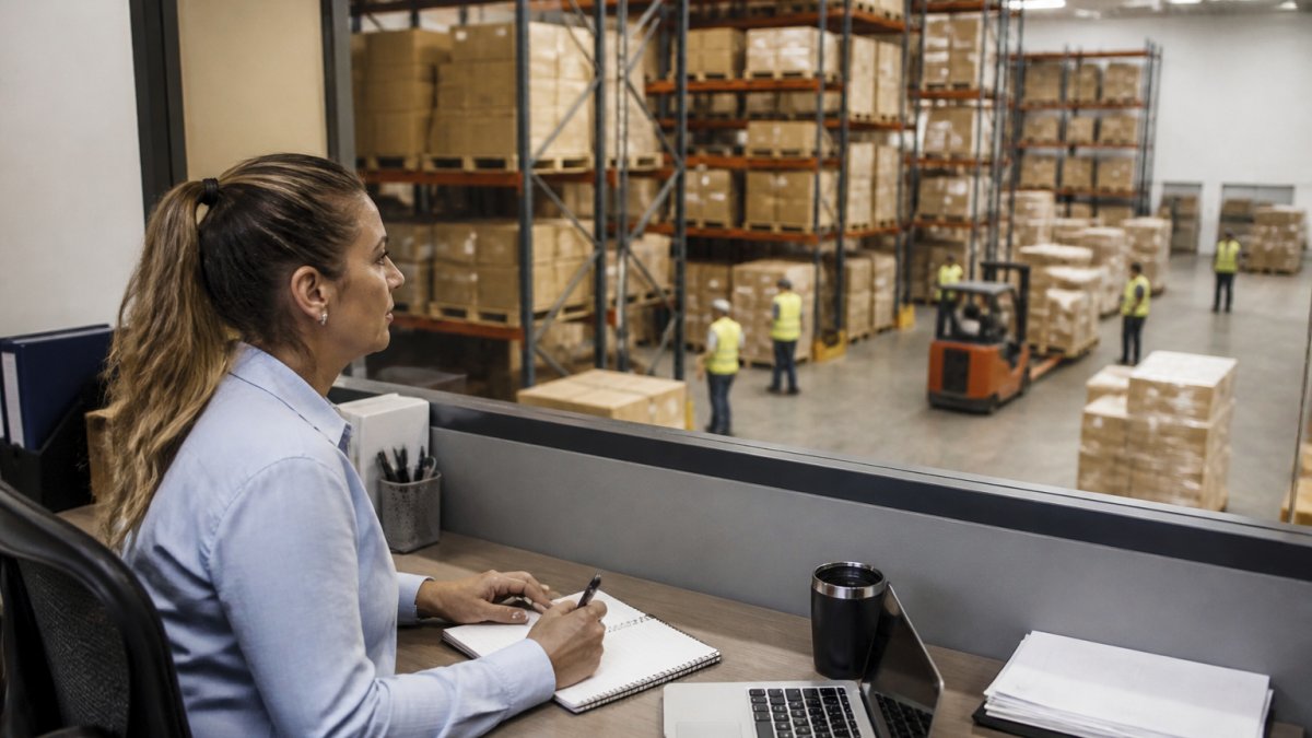 Operations manager at desk reviewing real-time inventory dashboards on multiple monitors with warehouse operations visible through window in background