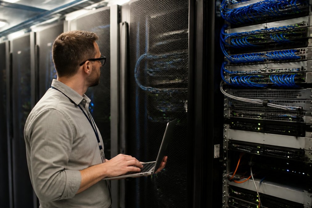 IT professional reviewing multiple server monitoring displays and network infrastructure diagrams in a modern data center with blue-lit equipment and cable management visible in the background