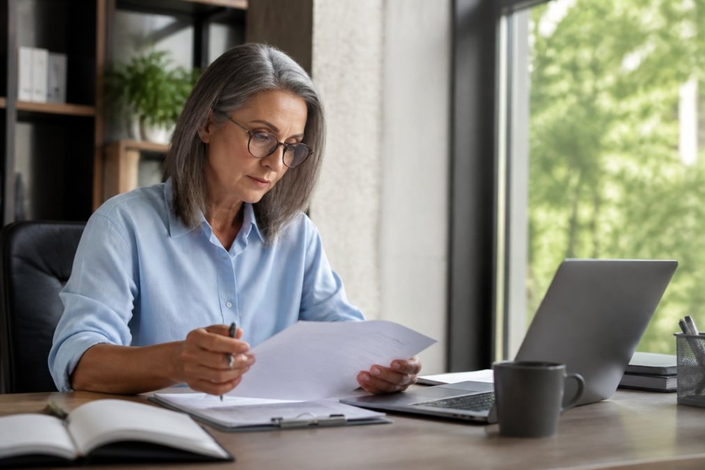 Experienced marketing director reviewing strategy notes and analytics on a laptop at a modern desk, with multiple monitors displaying performance dashboards and campaign metrics in a bright, modern office