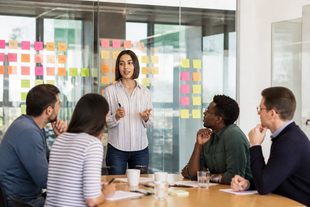A Scrum Master facilitating a planning session with a mid-sized software team in a bright meeting room, sticky notes arranged on a glass wall behind them