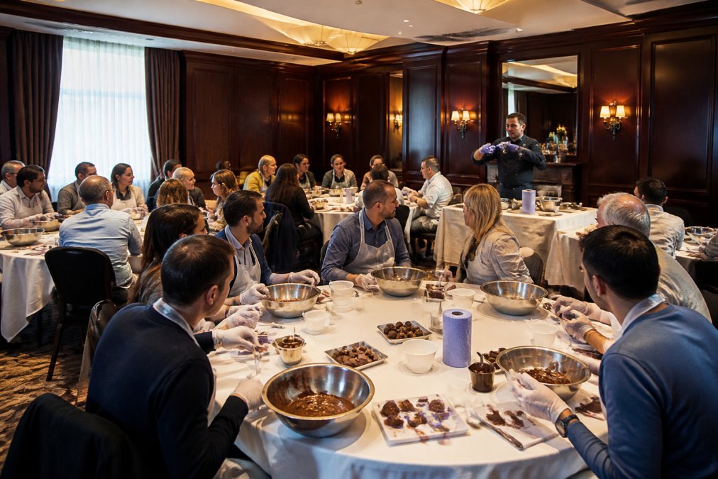 A large group of corporate employees seated at round tables during a hands-on chocolate-making workshop, actively engaged in tempering chocolate with a facilitator demonstrating at the front of a warmly lit conference room