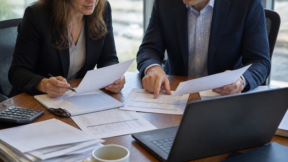 A property controller and an accounting manager reviewing printed close checklists and financial statements at a conference table, with a laptop open to accounting software in the foreground