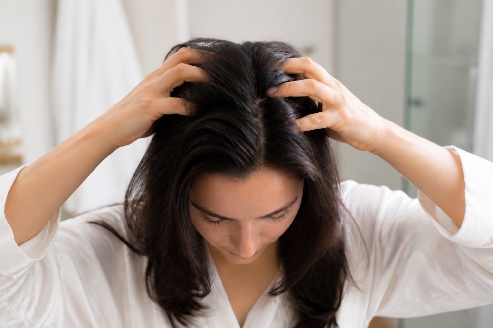 Close-up of a person's hands with fingertips placed on their scalp, demonstrating circular motion massage technique on dry hair in a bright bathroom