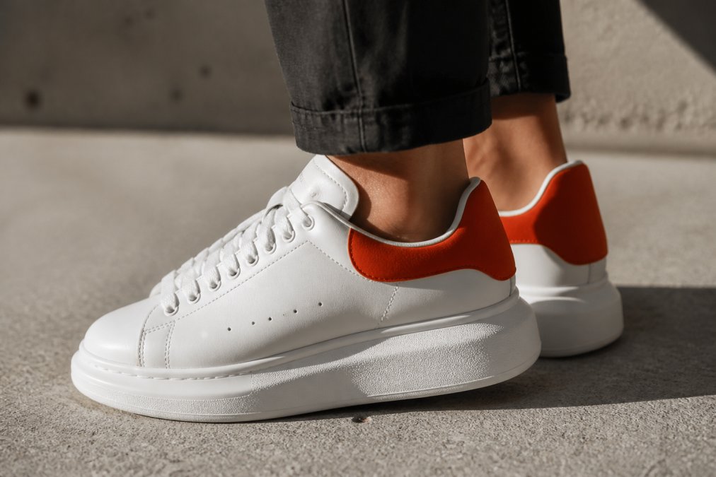 Primo piano di sneakers di lusso bianche con suola chunky e talloncino arancione sul retro, appoggiate su cemento chiaro con luce naturale laterale